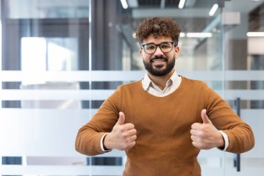 Modern ofis içinde, gözlüklü ve V yakalı süveter giyen, kendinden emin genç ve başarılı bir iş adamı iki eliyle olumlu bir onay hareketi gösteriyor.