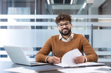 Genç profesyonel bir adam belgeleri gözden geçirir ve masasında notlar alır. Parlak, modern bir ofiste, yoğun bir iş gününde izleyiciye güvenle gülümserken.