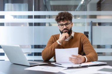 Çağdaş bir ofis masasında mali raporları ve yasal belgeleri inceleyen genç işadamı, çalışma gününde odaklandığını ve konsantre olduğunu gösteriyor.