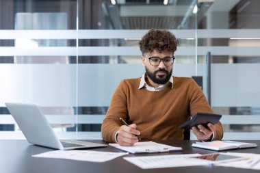Genç adam mali raporları ve hesap makinesiyle rakamları analiz ediyor, modern ofis ortamında bir masada otururken iş finans ve muhasebe üzerinde çalışıyor.