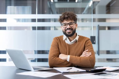 Gözlüklü ve kahverengi kazaklı bir işadamı modern bir ofiste bir masada oturuyor, gülümsüyor ve kameraya bakıyor, profesyonelliği ve başarıyı yansıtıyor.