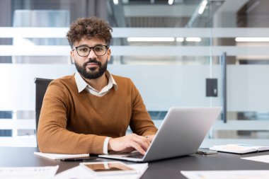 Masasında oturan gözlüklü ve kıvırcık saçlı genç profesyonel bir adam dizüstü bilgisayara bakıyor ve kameraya bakıyor. Şirket ortamında üretkenlik ve teknolojiyi somutlaştırıyor.
