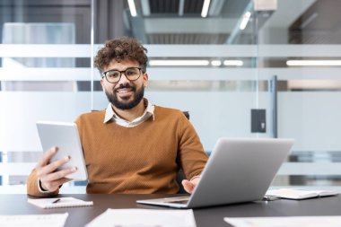 Kıvırcık saçlı ve gözlüklü genç adam bir ofis masasında oturuyor, elinde dijital tablet tutarken gülümsüyor ve bir dizüstü bilgisayar üzerinde çalışıyor, modern iş iletişimini ve teknolojisini temsil ediyor.