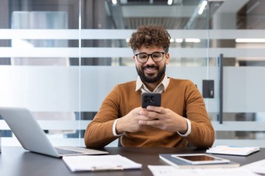 Akıllı bir telefon tutan genç işadamı, modern bir ofiste masasında otururken aktif olarak cep telefonu teknolojisiyle uğraşıyor, internet bağlantısının ve iş yerindeki iletişiminin tadını çıkarıyor.