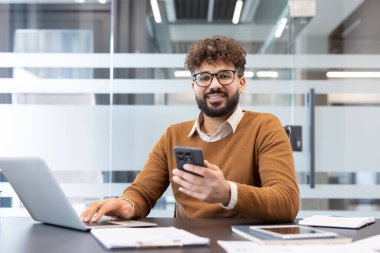 Sakallı genç işadamı dizüstü bilgisayarla çalışırken kameraya gülümsüyor ve masasında verimliliği ve modern iletişimi temsil eden akıllı telefonu tutuyor.