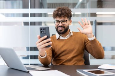 Cep telefonundaki video konferansında el sallayan kıvırcık saçlı ve gözlüklü genç işadamı, çağdaş ofis ortamında bir masada oturuyor.