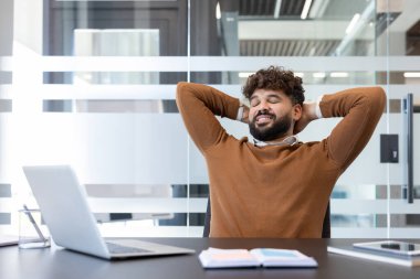 Genç işadamı, elleri masanın arkasında rahat bir şekilde huzurlu bir anın tadını çıkarıyor ve yanında bir dizüstü bilgisayarla işe ara veriyor.