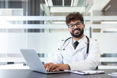 Genç Hintli doktor beyaz önlük ve steteskop giyiyor. Modern bir hastane masasında bilgisayarla çalışırken gülümsüyor. Tıbbi teknolojiyi ve ulaşılabilir sağlık hizmetlerini temsil ediyor.