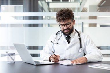 Önlüklü ve gözlüklü genç erkek doktor, bir klinik masasında oturuyor. Hasta sağlık raporlarını yazarken gülümsüyor. Modern bir muayenehanedeki dizüstü bilgisayarın yanında.