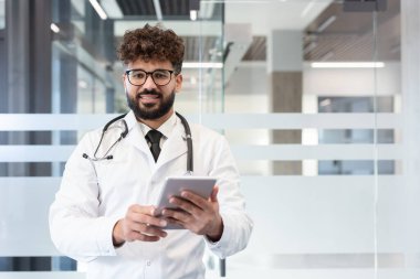 Beyaz önlüklü ve steteskoplu genç doktor gülümsüyor, hasta bakımı ve veri yönetimi için dijital tablet tutuyor, modern bir klinik veya hastane ortamında duruyor.
