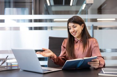 Dizüstü bilgisayardaki bir video konferansı sırasında gülümseyen ve konuşan genç bir iş kadını elinde bir kalem ve defter, modern bir ofiste uzaktan iş görüşmesi.