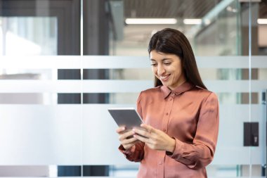 Parlak, modern bir ofiste gülümseyen ve tablet kullanan profesyonel iş kadını, dokunmatik ekran uygulamaları ve projeleri ve iletişimi yönetmek için dijital araçlarla güvenle çalışıyor