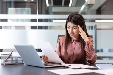 Belge tutan endişeli bir ifadeyle genç bir iş kadını, başağrısı çekiyor ve modern ofis çalışma alanındaki çok sayıda görev ve sıkı teslim tarihleri nedeniyle bunalmış hissediyor.