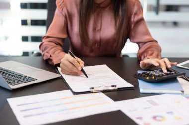 Kadınlar hesap makinesi kullanarak mali verileri hesaplayarak ve iş belgelerini yazarak, bir ofis masasında dizüstü bilgisayar ve çeşitli evrak işleriyle uğraşarak ellerini uzatıyorlar.