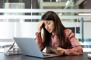 Kadın ofis masasında stresli ve endişeli hissediyor, kredi kartı tutarken finansal sorunlar yaşıyor ve online ödemeler veya bankacılık için dizüstü bilgisayar kullanıyor