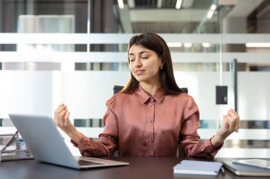 Modern ofis masasında dikkatli nefes alma pratiği yapan genç bir iş kadını, odaklanmayı, dengeyi ve zihinsel refahı artırmak için sakin olmak ve iş stresini azaltmak.