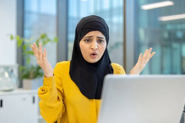 İnançsızlık ve hayal kırıklığını ifade eden tesettürlü Müslüman iş kadını, modern ofis ortamında dizüstü bilgisayara bakarken el kaldırıyor.