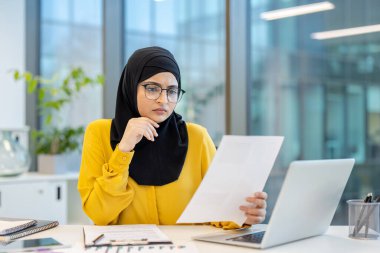 Çağdaş bir ofiste aktif olarak çalışan Müslüman bir iş kadını masasında dizüstü bilgisayar ve çeşitli ofis malzemeleriyle evrak ve belgeleri incelerken konsantre oluyor.
