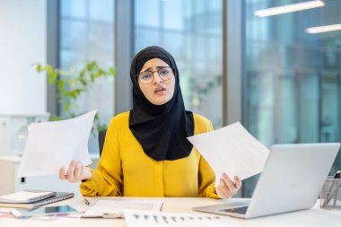 Başörtüsü takan ve gözlük takan Müslüman bir iş kadını bir yığın belgeyi yönetirken bir yandan da modern ofis masasında dizüstü bilgisayar kullanıyor.