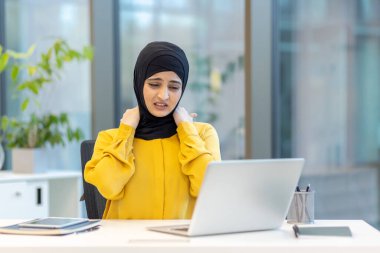 Müslüman bir kadın, ofis masasında dizüstü bilgisayarı üzerinde çalışırken keskin boyun ağrısı ve rahatsızlık yaşıyor. İşle ilgili stresi ve ergonomik sorunları vurguluyor.