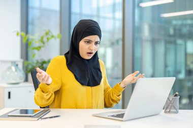 Başörtüsü içindeki Müslüman iş kadını, modern ofis ortamında çalışırken dizüstü bilgisayarındaki bir sorundan kaynaklanan kafa karışıklığı ve bitkinliği yaşıyor.