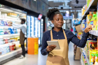 Önlüklü ve kareli gömlekli Afro-Amerikalı genç bir kadın market raflarındaki ürünleri kontrol etmek ve organize etmek için dijital tablet kullanıyor, envanter yönetimi ve verimli stoklama üzerine odaklanıyor.