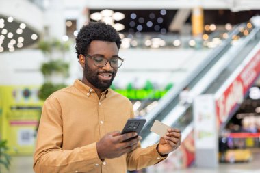 Elinde kredi kartı ve akıllı telefon olan gülümseyen Afrikalı bir adam, aydınlık bir alışveriş merkezinde dururken online alışveriş veya mobil bankacılık ile meşgul oluyor.