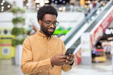 Alışveriş merkezinde gözlük takan gülümseyen bir adam, elinde akıllı bir telefonla ekranına bakıyor, arkadaşlarıyla bağ kuruyor ve çevrimiçi iletişimden keyif alıyor.