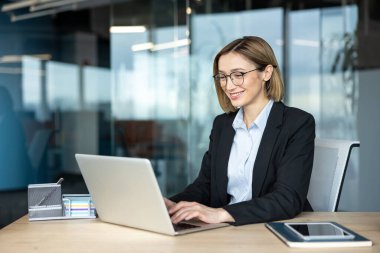 Profesyonel iş kadını dizüstü bilgisayarda gülümseyip daktilo kullanıyor, başarılı ve üretken bir çevreyi yansıtan cam duvarlı bir ofis ortamında çalışmalarını yönetiyor.