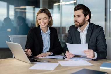 Modern bir ofiste birlikte çalışan iki farklı iş adamı. Bir kadın elinde belgelerle dizüstü bilgisayarı işaret ediyor. Proje detayları ve stratejileri tartışıyorlar.