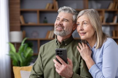 Üst düzey çift oturma odalarında modern teknolojinin tadını çıkarıyorlar, akıllı telefon kullanıyorlar ve gülümseyen yüzlerle başlarını çeviriyorlar, birlikteliği ve bağlantıyı kucaklıyorlar.