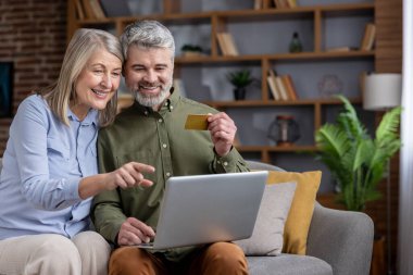 Oturma odasındaki kanepede oturan mutlu çift, gülümseyip dizüstü bilgisayara bakarken kredi kartıyla online ödeme yapıyorlar, modern dijital kolaylığın tadını çıkarıyorlar.