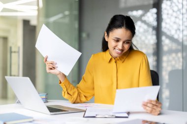 Mutlu iş kadını ofis masasında önemli belgeleri ve evrakları inceliyor, bir dizüstü bilgisayarla çalışıyor ve gülümsüyor, kariyerindeki başarı ve profesyonelliği temsil ediyor.