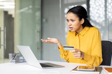 Online alışveriş sırasında kredi kartı ödemesi sorunu ya da hatası yaşayan kafası karışmış bir kadın, ofis masasında dizüstü bilgisayarla oturuyor ve elinde bir bankacılık belgesi tutuyor.