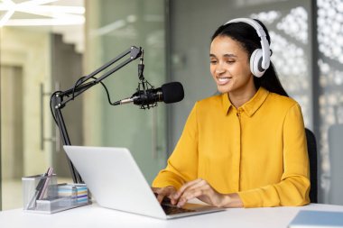 Genç mutlu içerik yaratıcısı, yayın medyası üretimi ve çevrimiçi iletişimi temsil eden mikrofon, kulaklık ve dizüstü bilgisayar kullanarak bir podcast kaydediyor.