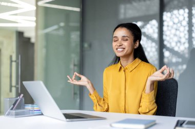 Genç iş kadını gözlerini kapatıp gülümsüyor ve Lotus Mudra parmaklarıyla meditasyon yapıyor. Stres atmak ve sağlığını arttırmak için modern ofis masasında poz veriyor.
