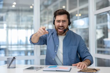Erkek sakallı çağrı merkezi temsilcisi, kulaklık takıyor, agresif bir şekilde bağırıyor ve ofis ortamında bir masada otururken öne doğru işaret ediyor, hayal kırıklığını ve çatışmayı ifade ediyor.