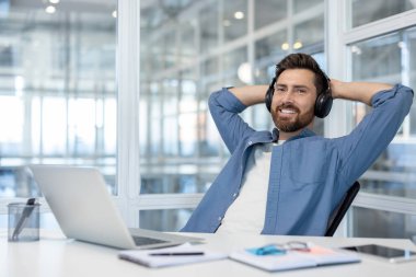 Gülümseyen bir adam, kulaklıkla müzik dinlerken rahatlamanın keyfini çıkarıyor, modern bir çalışma alanında başarılı bir iş molası sırasında koltuğuna yaslanıyor.