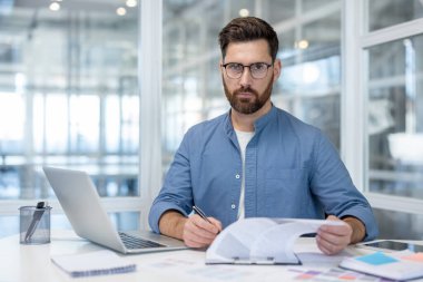 Profesyonel bir adam gözlüklü ve masasında oturan mavi gömlekli, elinde belgeler, notlar tutan, ve parlak bir ofis ortamında çalışırken bir dizüstü bilgisayarının açık olması.