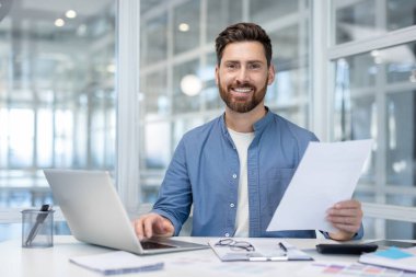Ofis masasında çalışan gülümseyen işadamı, elinde bir belge tutuyor ve bir dizüstü bilgisayar kullanıyor, başarı, üretkenlik ve profesyonel başarı kavramlarını temsil ediyor.