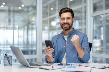 Gülümseyen adam heyecanlanıyor ve yumruk pompası yapıyor, zaferi veya başarıyı cep telefonundan aldığı iyi haberle modern ofis ortamında kutluyor.