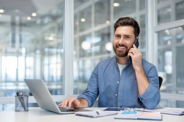 Kendine güvenen sakallı adam modern ofiste masa başında oturuyor, dizüstü bilgisayarda yazıyor ve mutlu bir şekilde cep telefonuyla konuşuyor, uzaktan çalışmayı ve profesyonel iletişimi temsil ediyor.