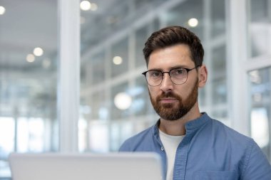 Gözlüklü ve sakallı bir adam kameraya bakıyor, modern ofis arka planında poz veriyor, profesyonelliği, odaklanmayı ve çalışırken kurumsal başarıyı yansıtıyor.