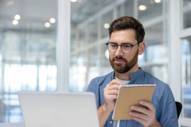 Gözlüklü girişimci veriye yoğunlaşırken aynı zamanda sarmal bir not defterine not alıyor, parlak ve modern bir ofiste dizüstü bilgisayarla iş görevlerine katılıyor.