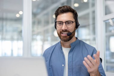 Gülümseyen erkek çağrı merkezi ajanı mikrofonlu bir kulaklık takıyor, bir dizüstü bilgisayar kullanıyor ve bir müşteriyle çevrimiçi video görüşmesi ya da tele pazarlama servisi aracılığıyla iletişim kurarken işaret ediyor.