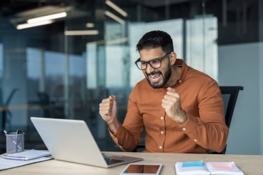 Heyecanlı Hintli adam dizüstü bilgisayara bakarken sevinçle yumruk atıyor ve bağırıyor, iş başarısını kutluyor ya da modern ofis ortamında online oyun kazanıyor.
