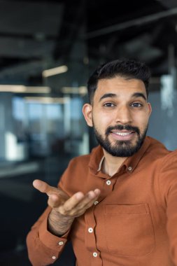 Kahverengi gömlekli genç sakallı bir adam video konferans sırasında iletişim kuruyor ve sunuyor. Ofis ortamında el hareketleriyle kameraya bakıyor.