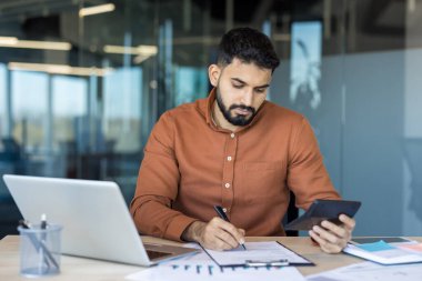 Genç bir finans uzmanı masada oturuyor, hesap makinesi kullanarak para sayıyor, belgeleri inceliyor, çizelgeleri ve grafikleri analiz ediyor ve modern ofis ortamında notlar alıyor.