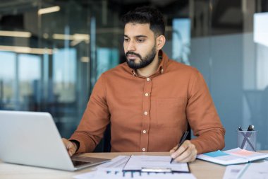 Kahverengi gömlekli Hintli bir adam, ofis masasına odaklanmış, bir dizüstü bilgisayarında yazı yazarken, dokümanlar hakkında notlar alıyor, çizelgeleri analiz ediyor ve iş görevlerini konsantrasyon ve amaçla planlıyordu.