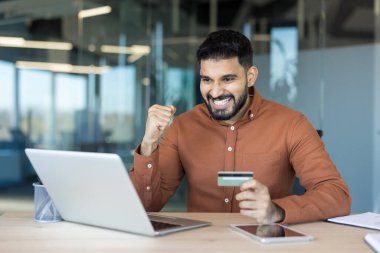 Gülümseyen genç adam başarılı bir online alımı kutluyor, bilgisayarının yanında bir kredi kartı tutarken güvenli bir e-ticaret alışverişi ve tamamlanmış ödemenin sevincini dile getiriyor.
